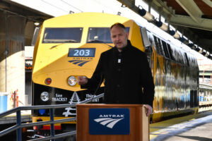 Man on podium in front of yellow and black locomotive parked on station platform