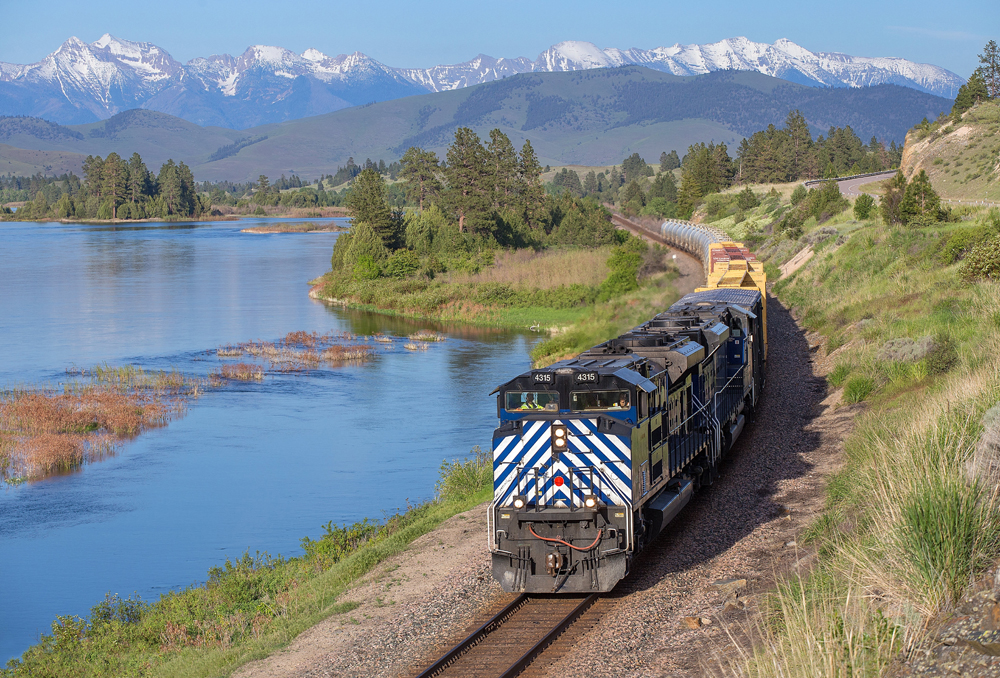 A blue Montana Rail Link train works alongside a river with mountains in the background.