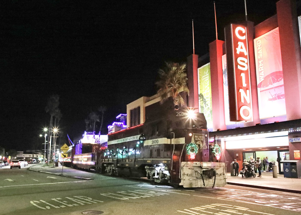 Maroon locomotive leads train with holiday lights down street at night