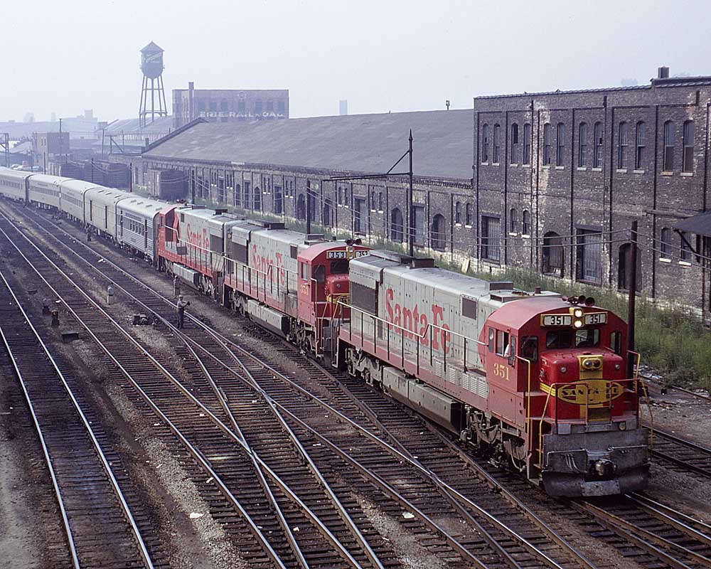 Three red-and-silver diesel locomotives pull a passenger train through an urban canyon