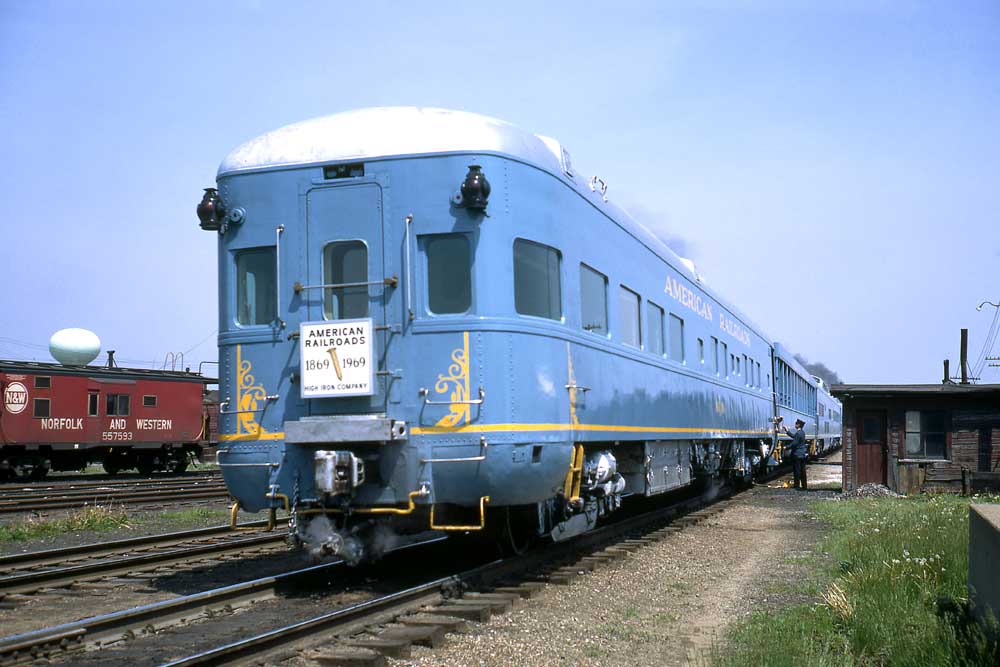 End of blue passenger train with sign highlighting railroad spikes