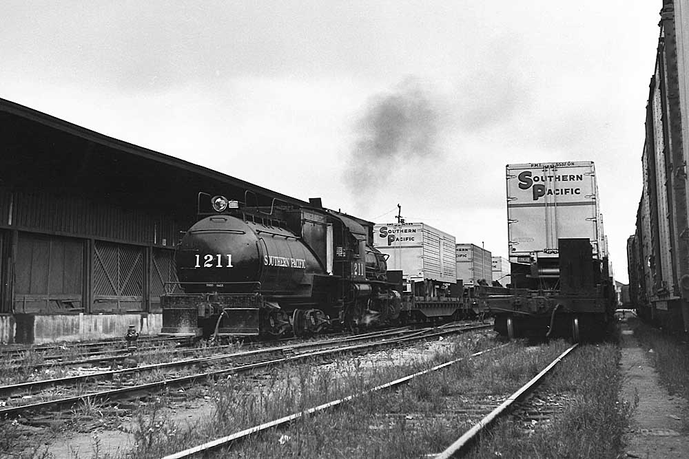 Southern Pacific freight trains: Steam locomotive pulls flat cars with trailers in yard alongside warehouse