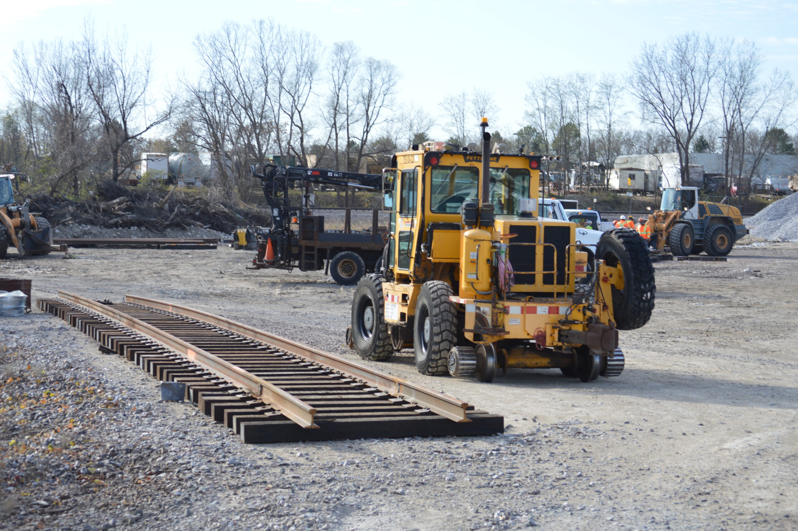 A piece of panel track lays in the staging yard next to a yellow construction tractor with rubber tires and rail wheels.