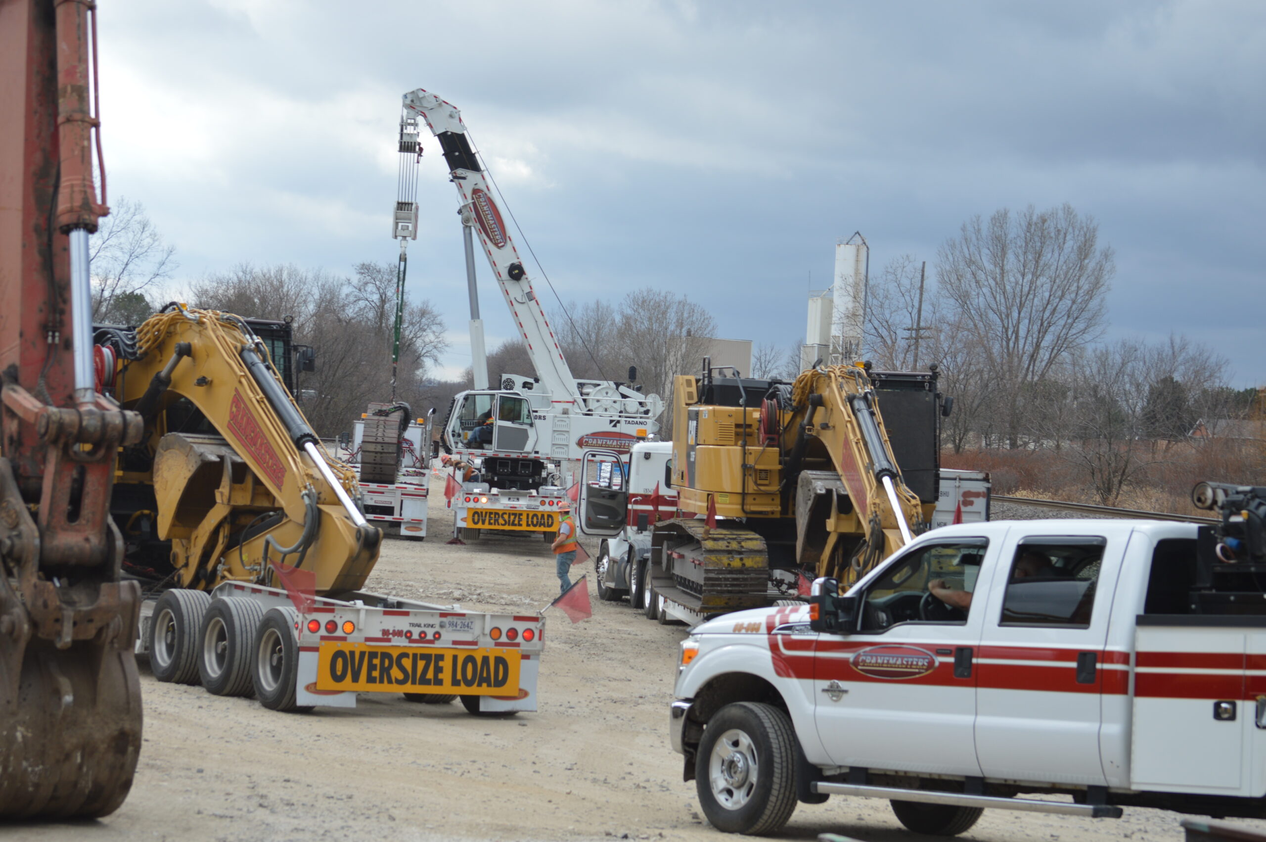 Pieces of construction equipment, including cranes and excavators, arrive on low-boy flatbed semi-trailers.