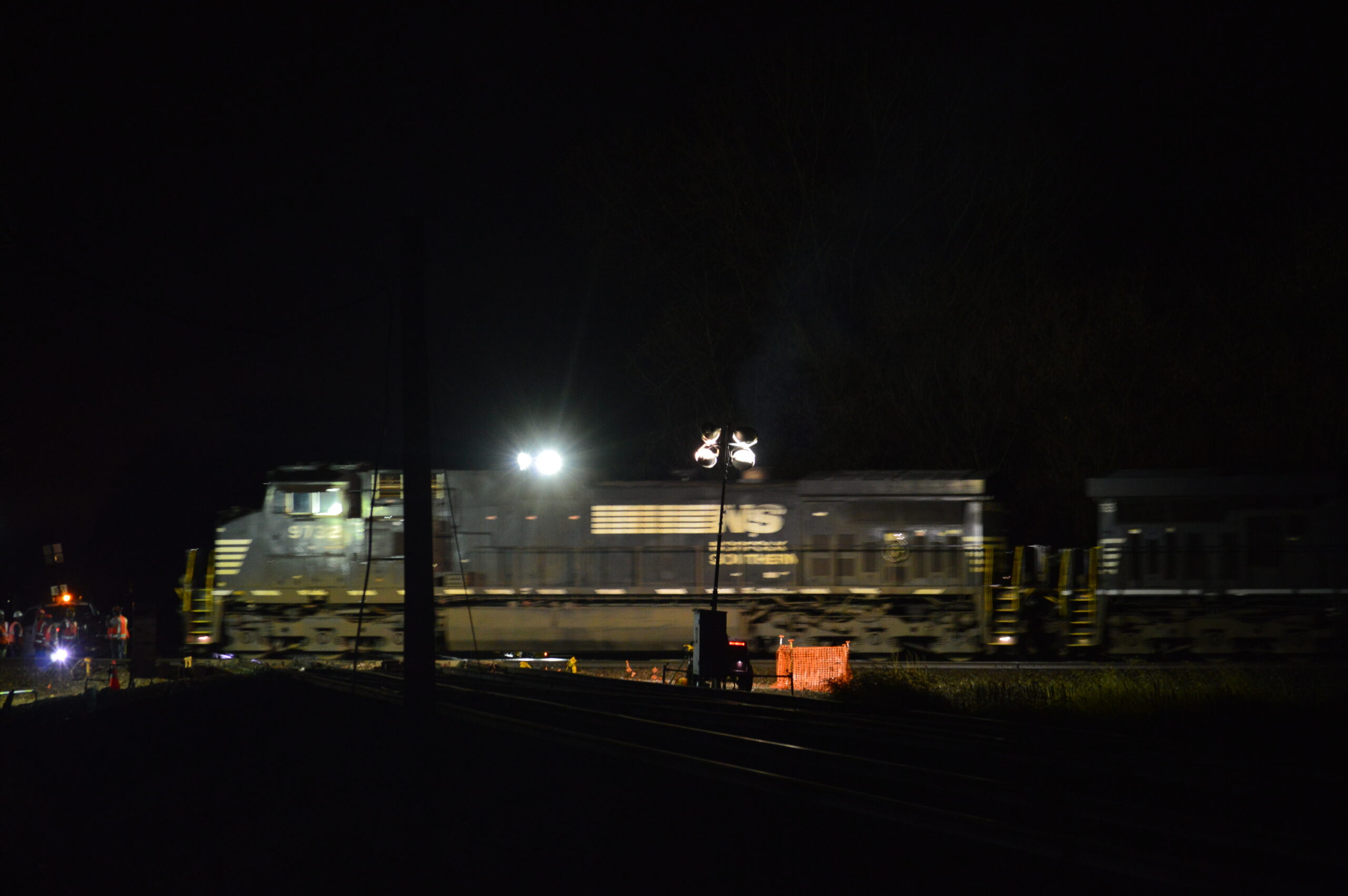 A black Norfolk Southern General Electric Dash 9 diesel is lighted by spotlights as it crosses the diamond in the dark.
