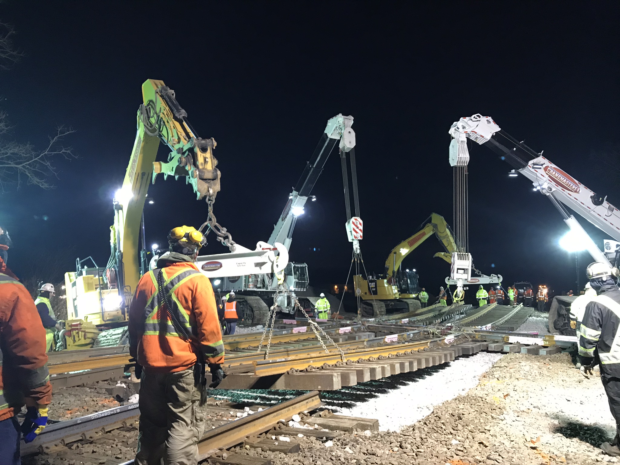 Four cranes place the new diamond on the roadbed as orange-vest clad crew members supervise.