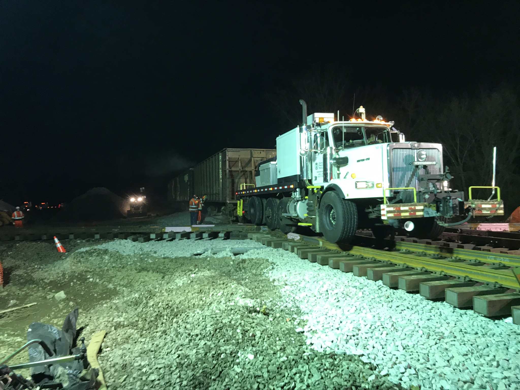 A flatbed truck with rail wheels positions ballast hoppers at the crossing.