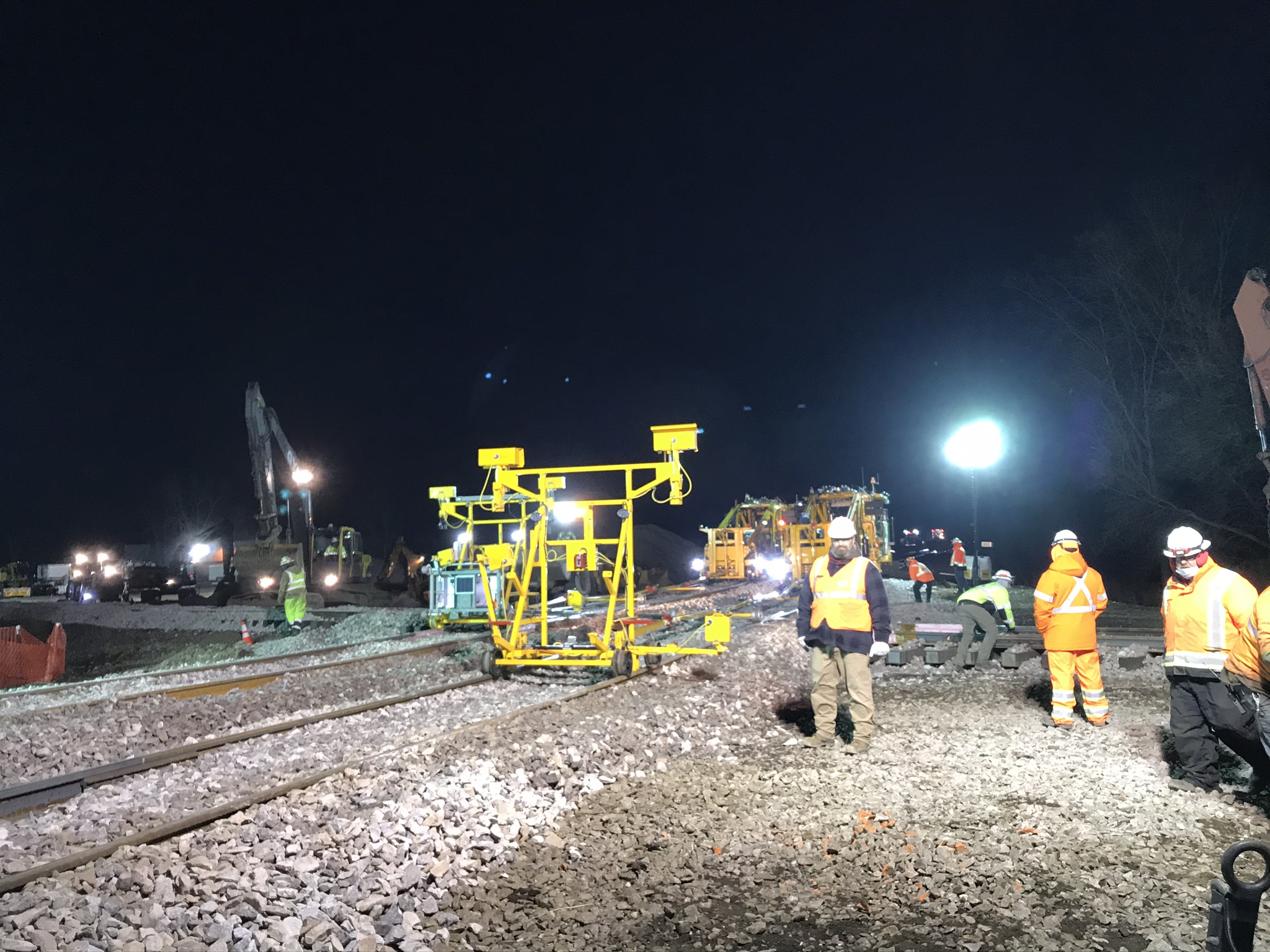 A yellow target frame rests on the northside Canadian Pacific track as orange-vest-clad workers supervise the work under spotlights.