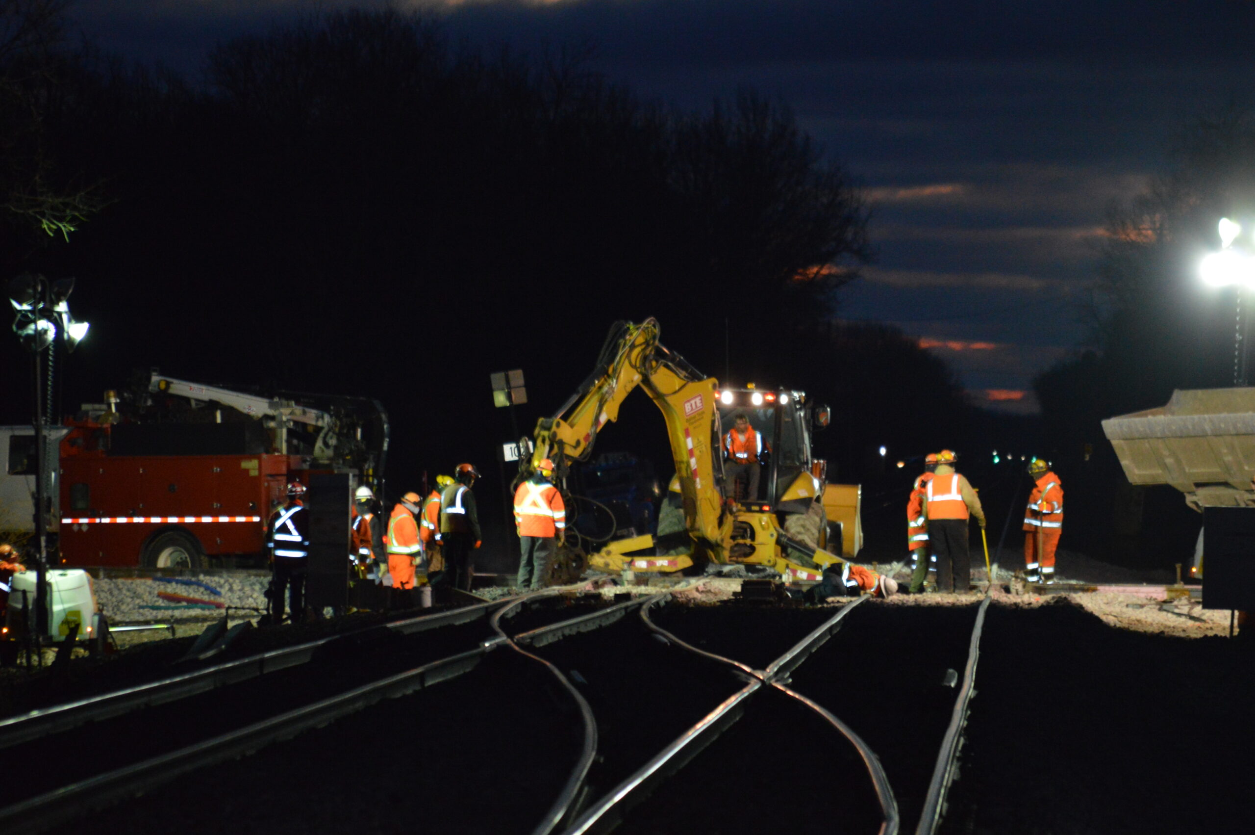 Spotlights highlight a yellow tamping machine attended by orange-vest-clad workers.