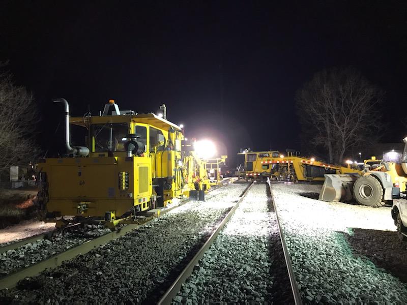 Yellow-painted construction equipment including ballast groomers and front-end loaders stand on the two-track Canadian Pacific main under spotlights.