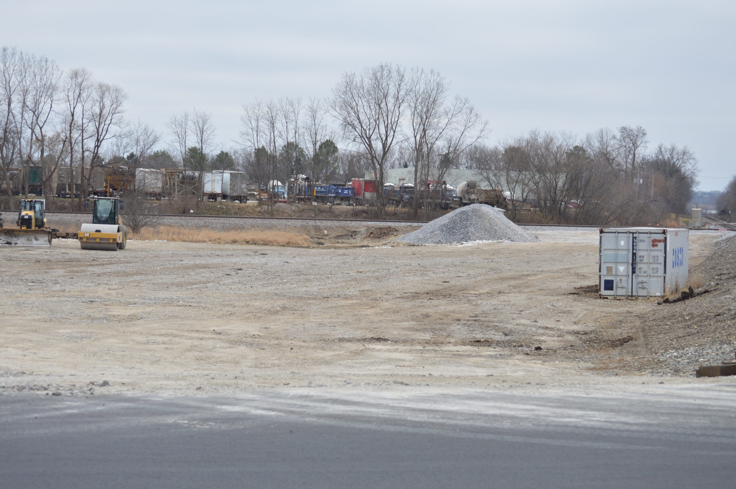 An empty staging yard with only a bulldozer and road roller, a large pile of gravel, and a gray construction container sitting in it on a gray day.