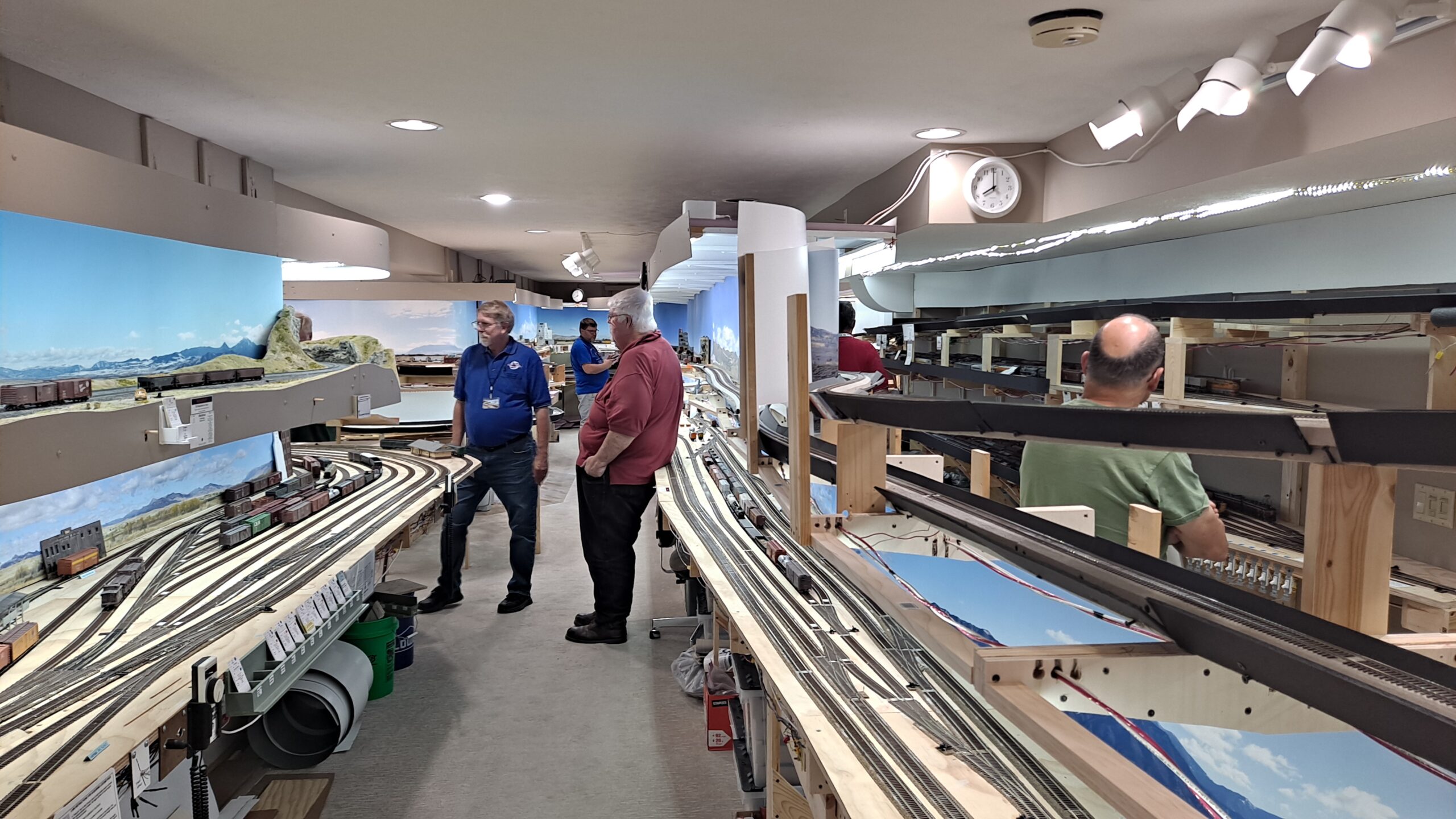 Model railroad benchwork on two levels runs along a long aisle as people mill around before an operating session.