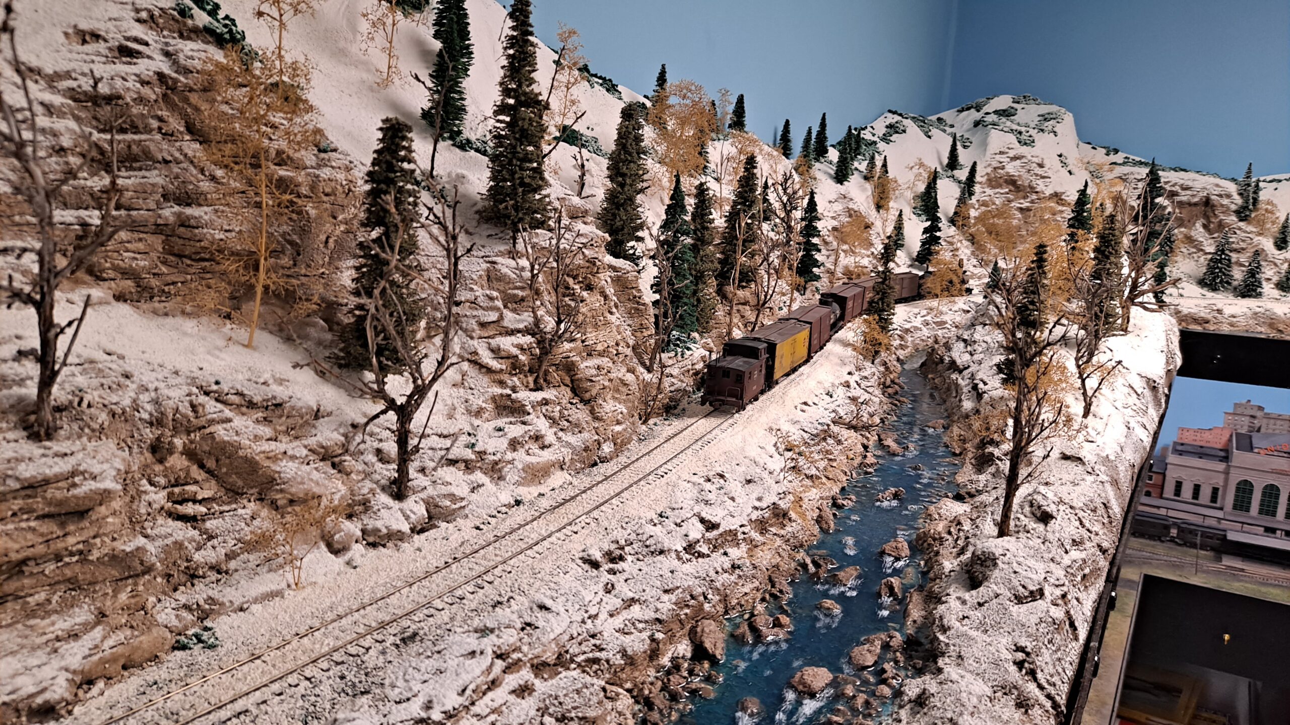 A steam-powered freight train runs through a snowy landscape at the bottom of a rocky canyon along a mountain stream.