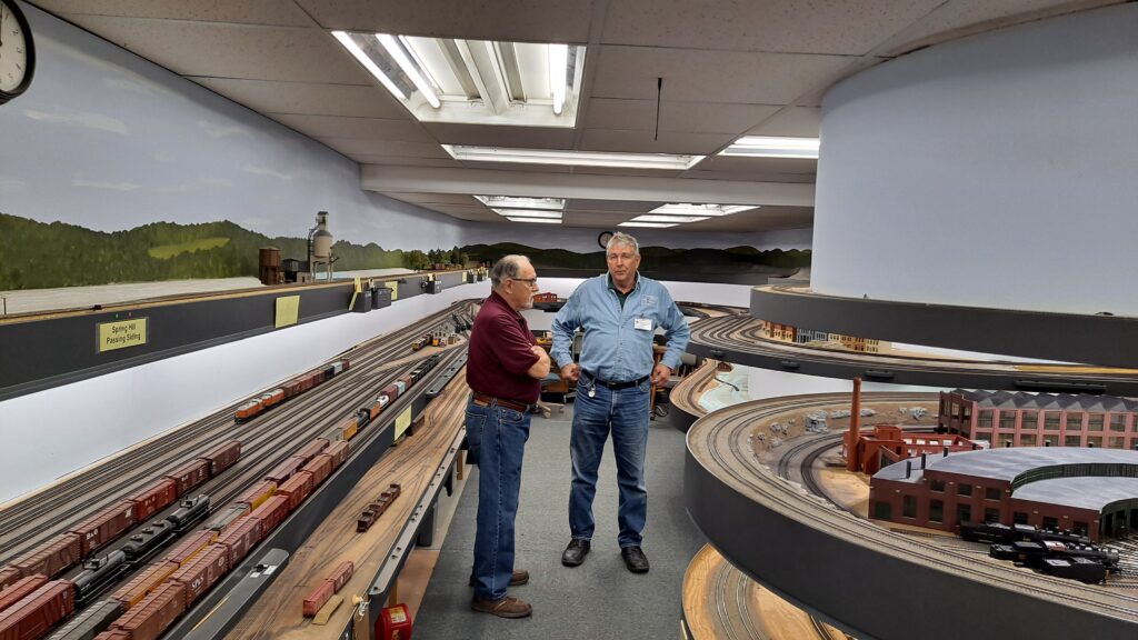 Two men talk at the intersection of two aisles of a double-deck model railroad.