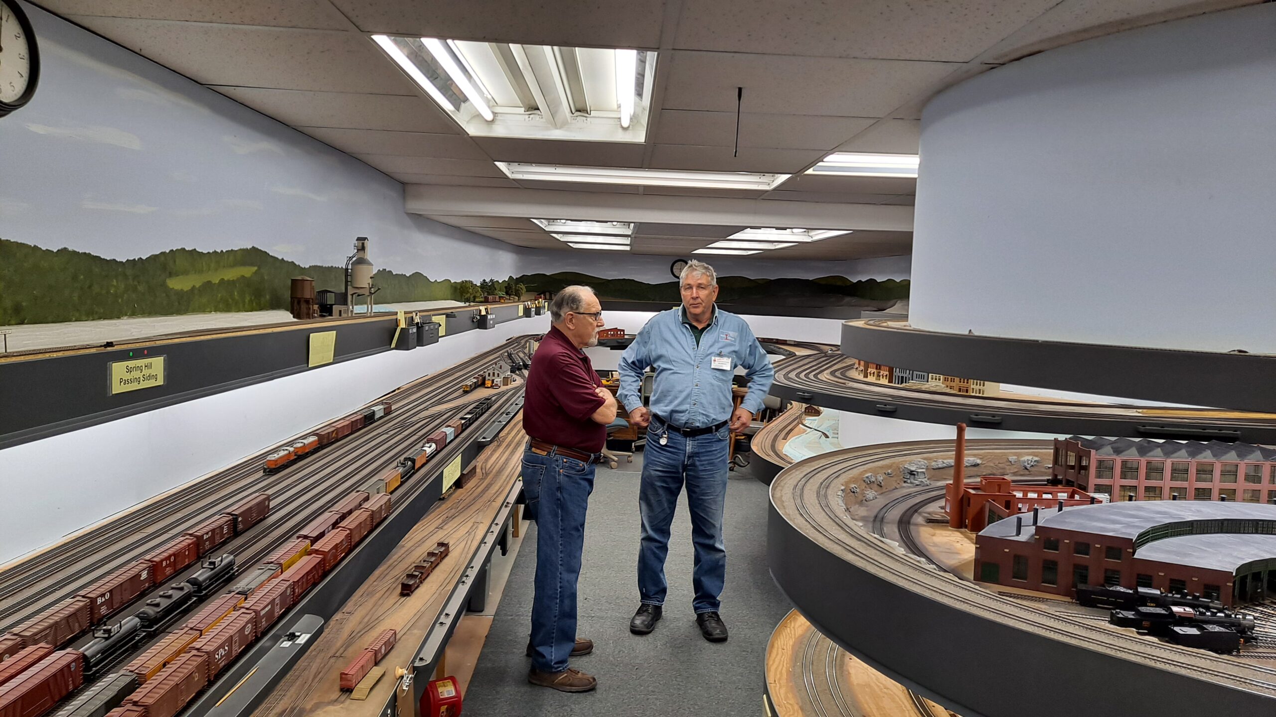 Two men talk at the intersection of two aisles of a double-deck model railroad.