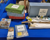Catalog, pamphlet, instruction book, three small, stacked boxes, one large blue gray box behind a sheet of Styrofoam with track, cork roadbed, foam brushes, paints, glues, and small tools.