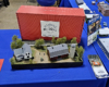 Diorama with farmhouse and barn with silo surrounded by trees with a single track running at the back of the scene. Red Build A World box is standing behind.