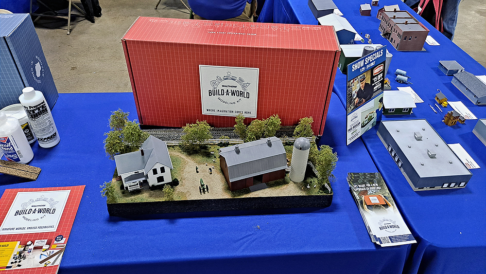 Diorama with farmhouse and barn with silo surrounded by trees with a single track running at the back of the scene. Red Build A World box is standing behind.