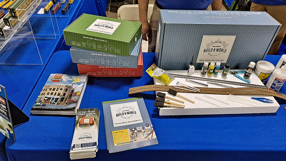 Catalog, pamphlet, instruction book, three small, stacked boxes, one large blue gray box behind a sheet of Styrofoam with track, cork roadbed, foam brushes, paints, glues, and small tools.