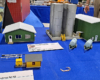 Green metal office building and garage with four tall silver tanks inside containment wall, two fertilizer tank trailers, yellow speeder shed with speeder, black and white striped crossing gates.