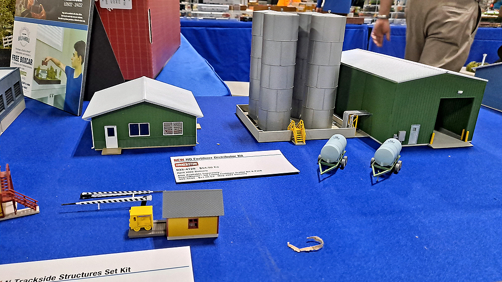 Green metal office building and garage with four tall silver tanks inside containment wall, two fertilizer tank trailers, yellow speeder shed with speeder, black and white striped crossing gates.