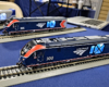 A blue passenger locomotive with red highlights sits on gray and black plastic track with another similar model in the background.