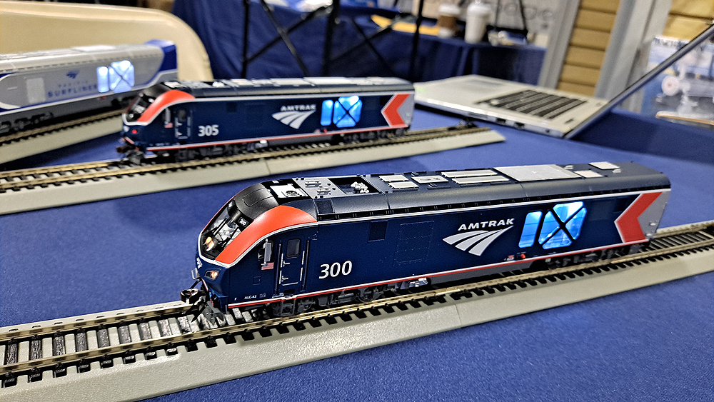A blue passenger locomotive with red highlights sits on gray and black plastic track with another similar model in the background.