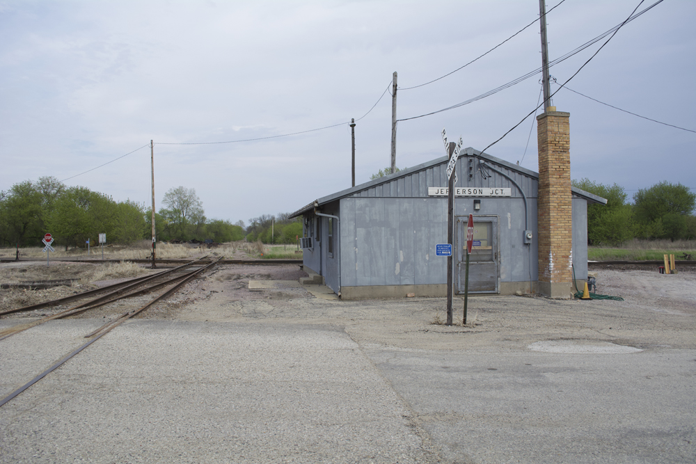 A gray metal railroad station in the southwest corner of at-grade railroad crossing.