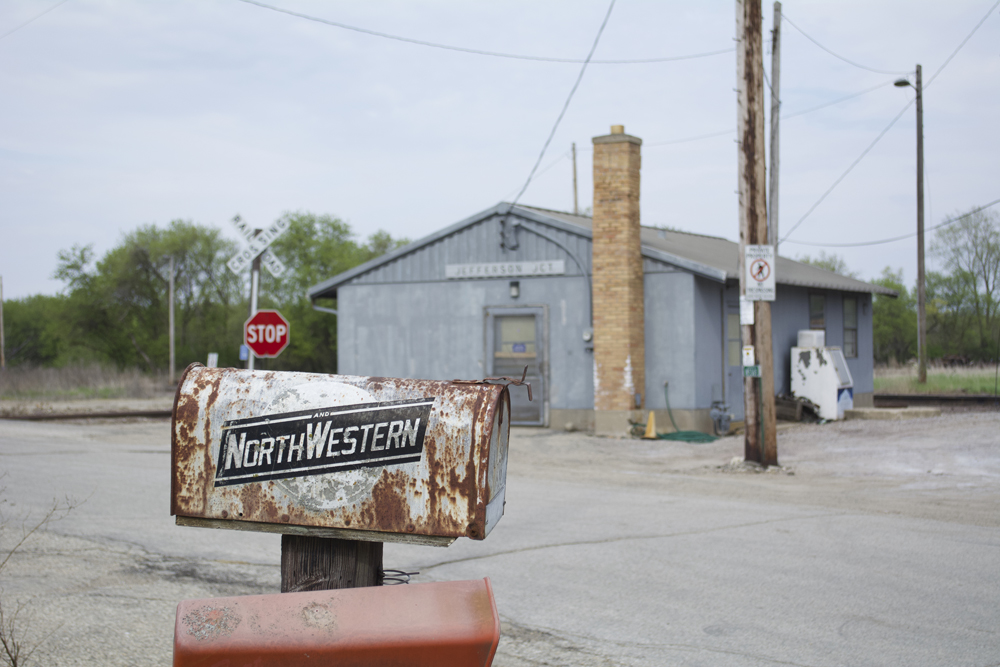 A rusty mailbox with worn Chicago & North Western herald on the side and a metal station building visible in background.