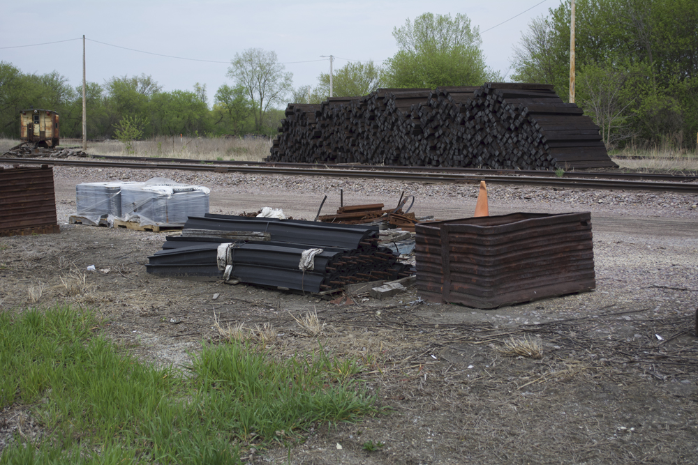 Spike kegs, joint bars, and metal storage containers sitting on gravel. In the background is a pile of new crossties.