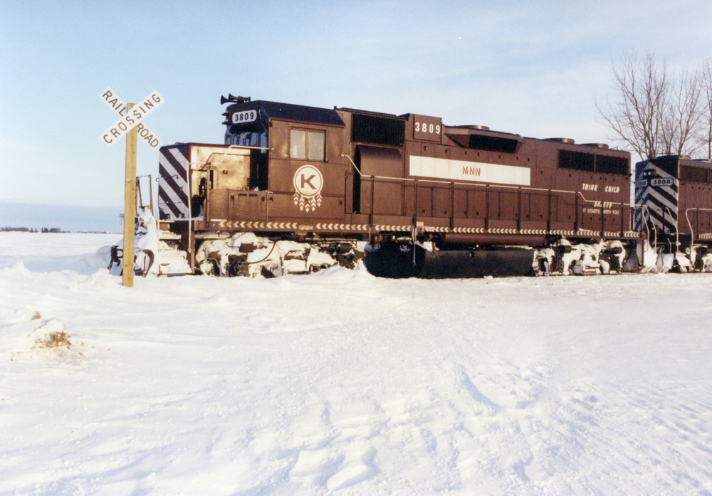 : In a snow-covered landscape, an Electro-Motive Division hood unit painted maroon with white graphics is parked by a railroad crossing buck attached to wood post.