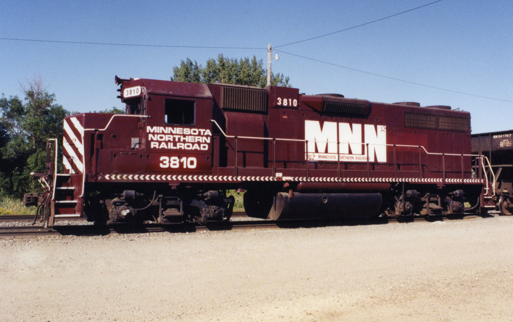 Photo of maroon EMD GP38 with white graphics pulling an open hopper on a sunny summer day.