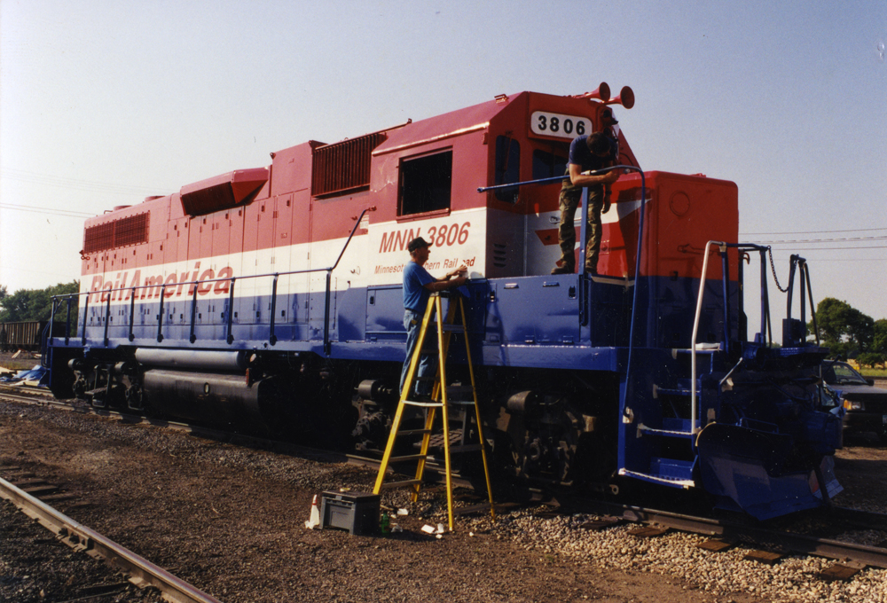 Workers apply paint and lettering to an EMD hood unit painted red, silver, and blue. 