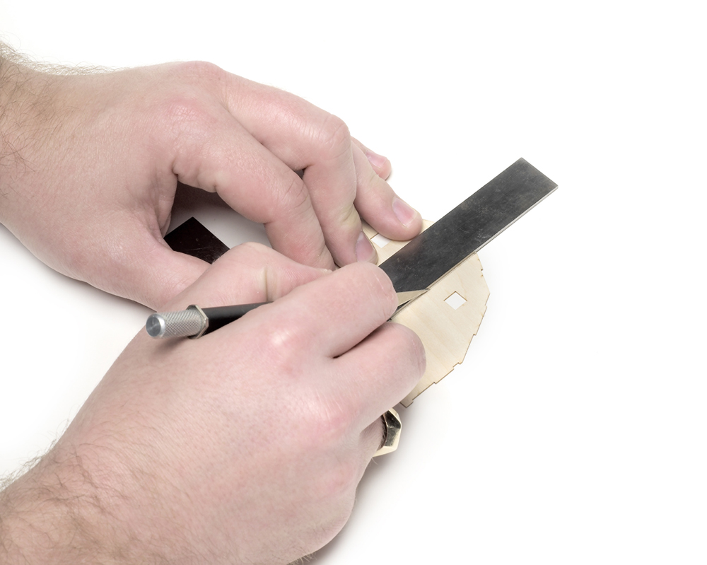 A hobby knife seated against a machine square cutting through a wood part.