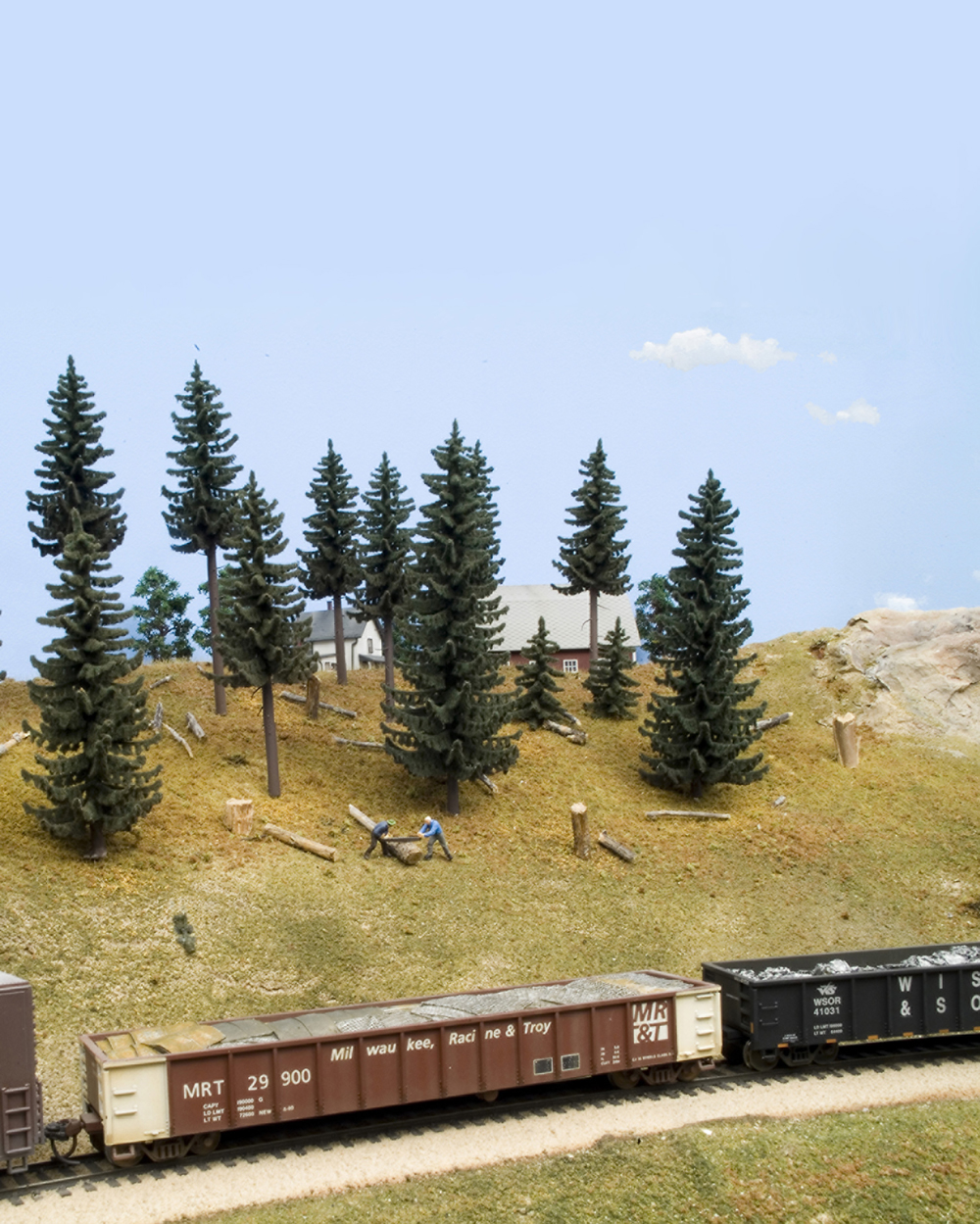 HO scale scene with gondola in foreground, trees behind track, and N scale structures in the distance.