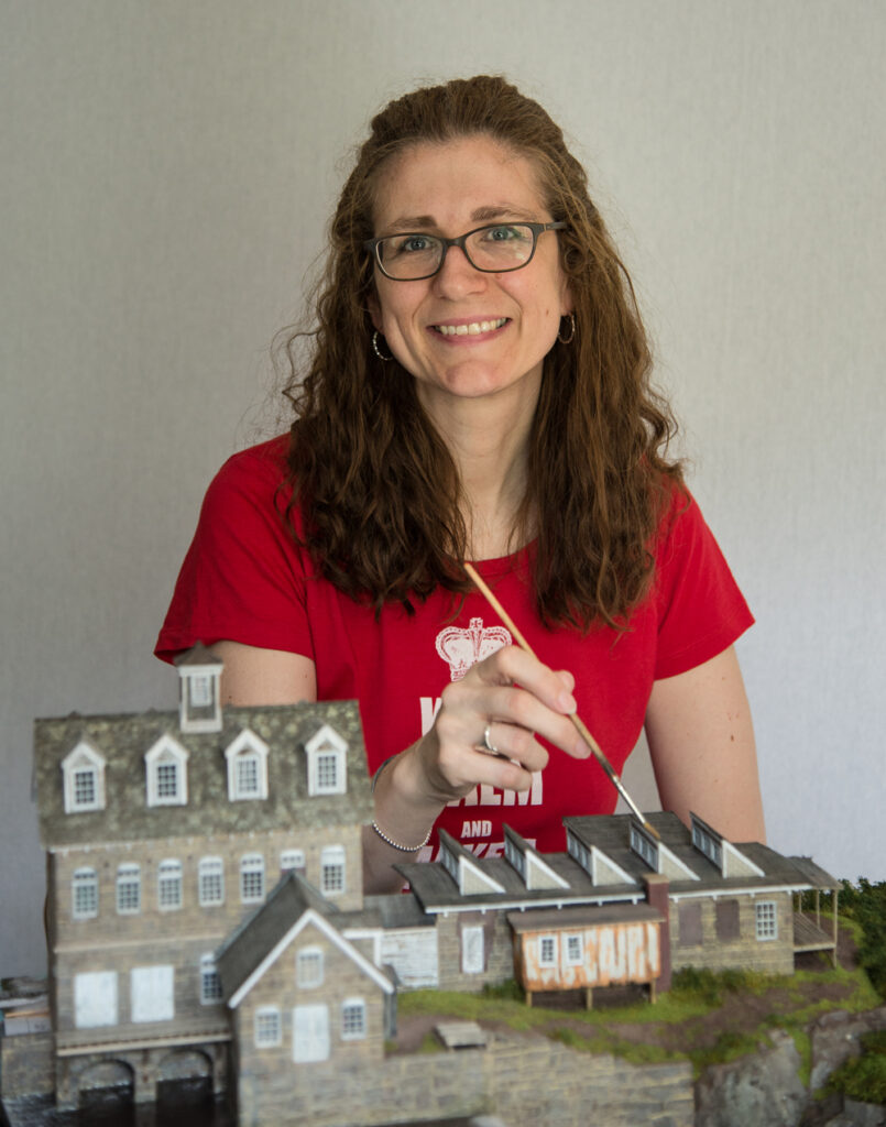 Kathy, in red T-shirt, poses with a paintbrush and several structures