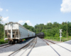 Derailed covered hopper and black tank car, part of another train, next to it on another track, just past a grade crossing