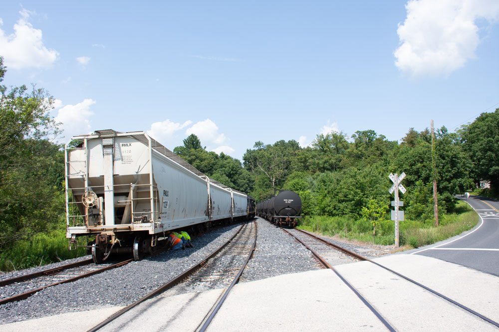 Derailed covered hopper and black tank car, part of another train, next to it on another track, just past a grade crossing