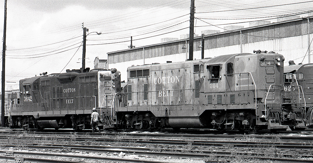 black and white photo of two locomotives couple on tracks next to building and a man with hard hat walking