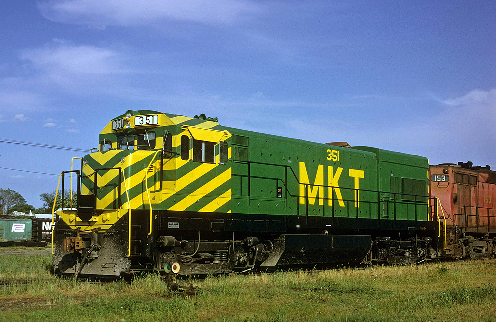 Locomotive with green body and green and yellow striped nose resting on grassy, overgrown tracks