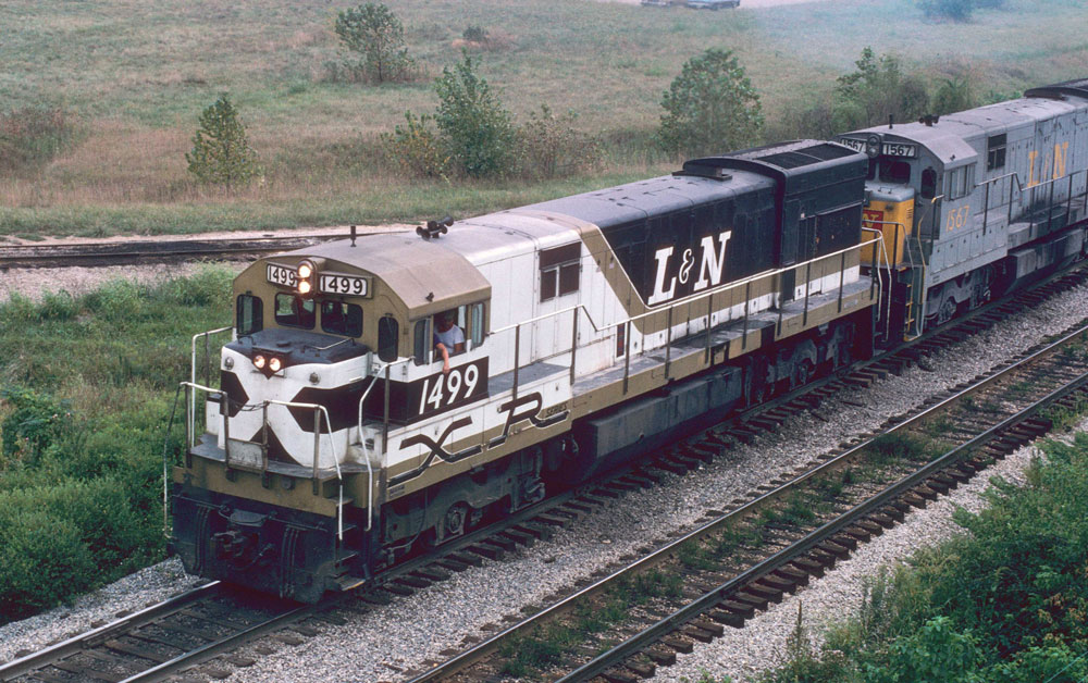 white, gold, brown, and black locomotive with people in cab with another locomotive coupled behind it