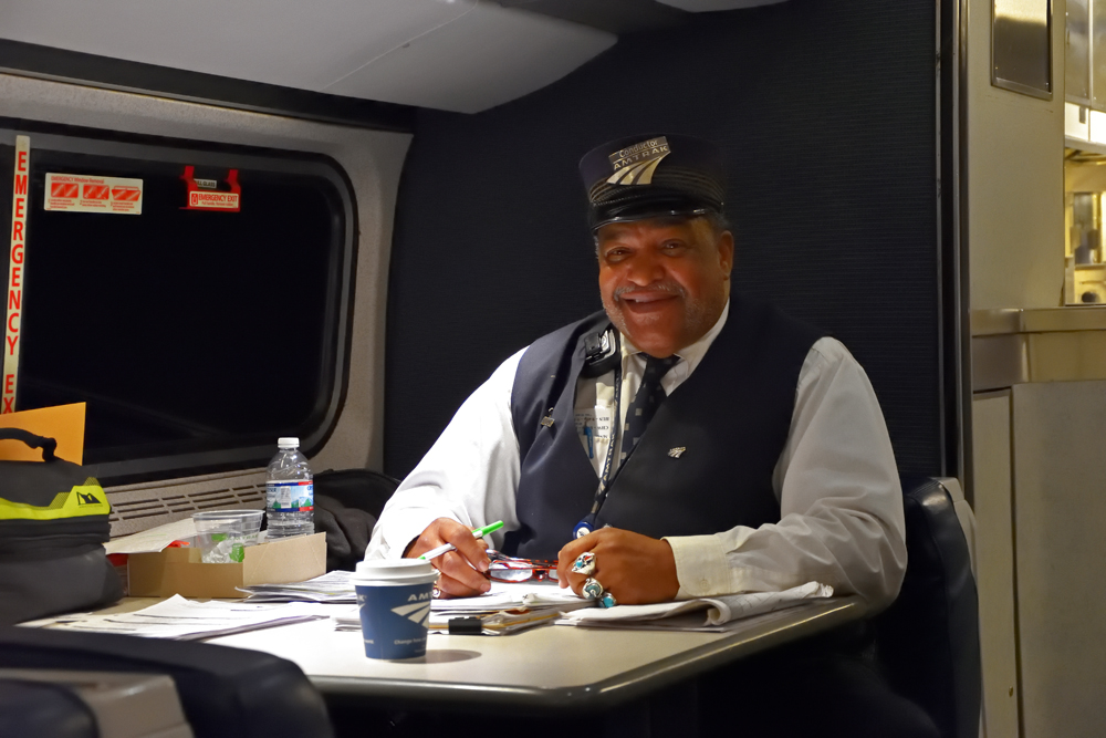 Man sitting at table in a passenger car surrounded by paperwork.