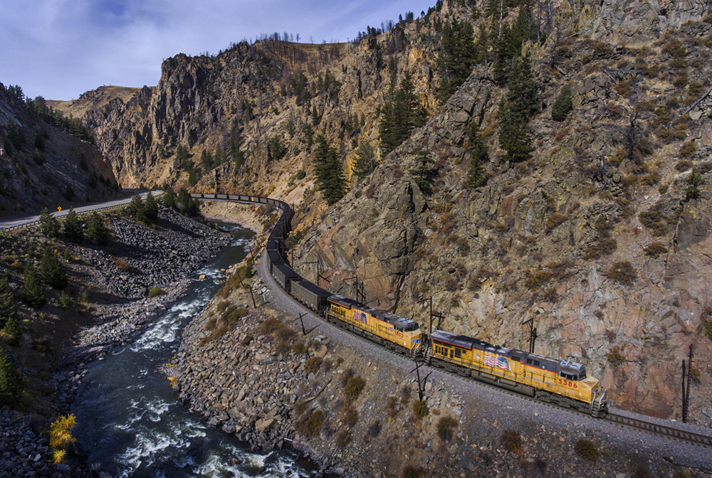 Train with two yellow diesels passes through S-curve parallel to river in rocky canyon.