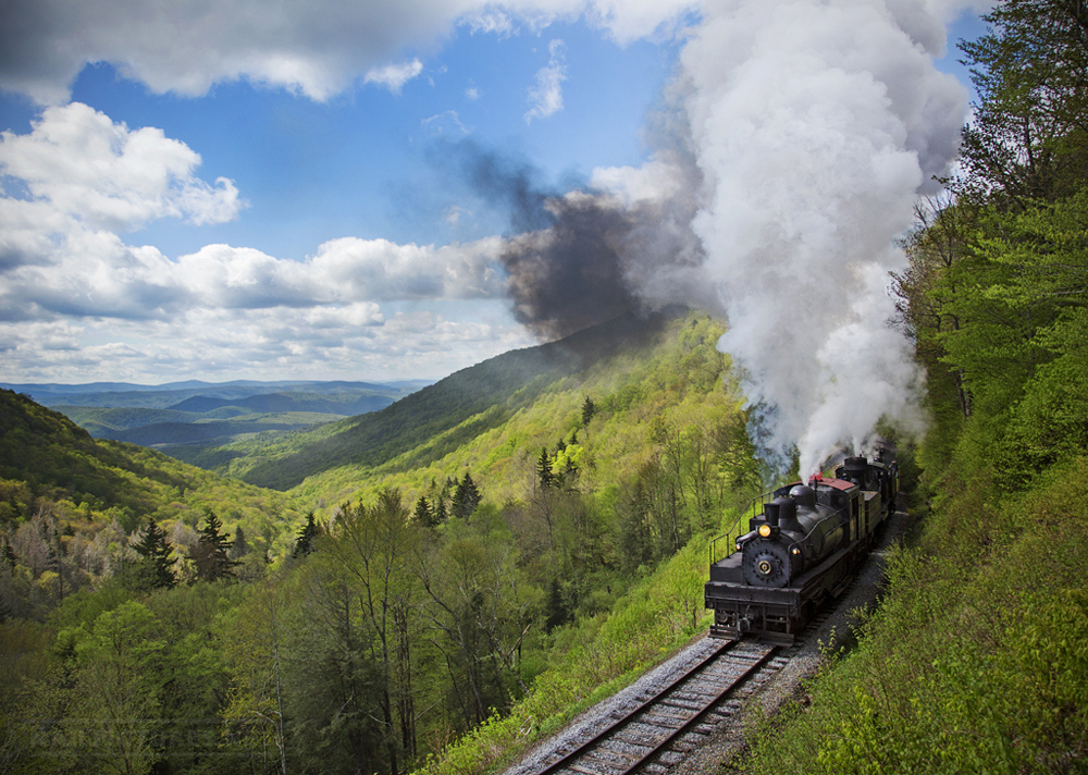 Black steam locomotive under plume of smoke at front right of image, passing through green mountains extending into the distance