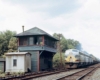 Streamlined diesel locomotives pull freight train past wooden interlocking tower