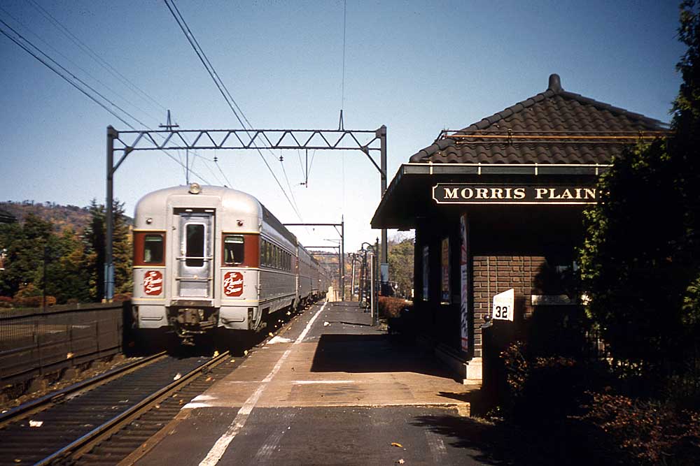 Rear of passenger train passes brick passenger station under catenary wires