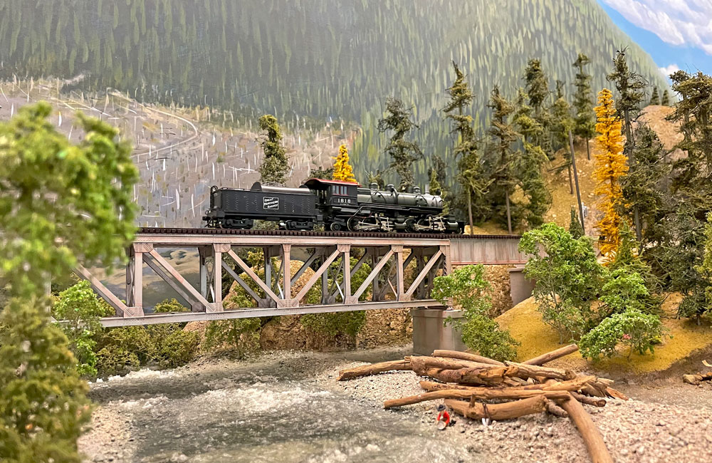 A Great Northern 2-6-6-2 steam locomotive crosses a steel deck truss bridge over a creek in wooded mountains.