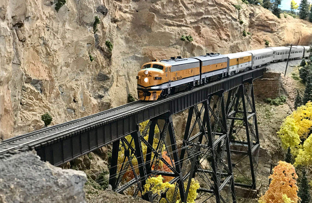 A gold-and-silver hood unit leads a passenger train onto a high steel trestle.
