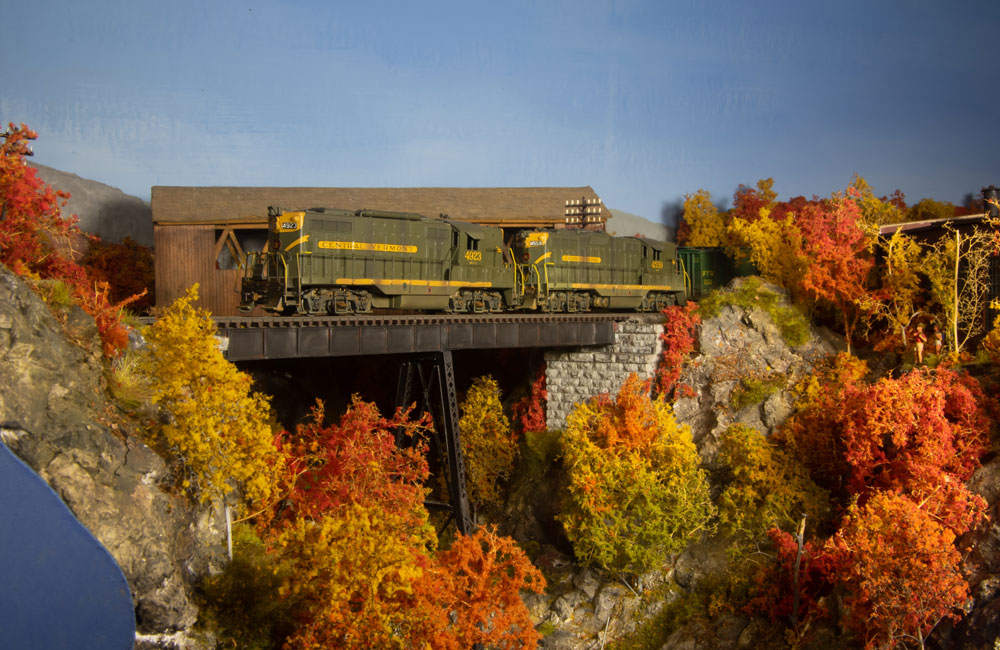 A pair of green-and-yellow Geeps step out onto a trestle amid brilliant fall foliage.
