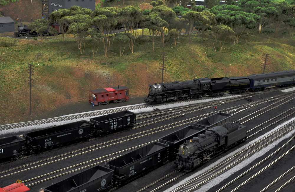 A black steam engine leads a mail train around the outskirts of a classification yard.